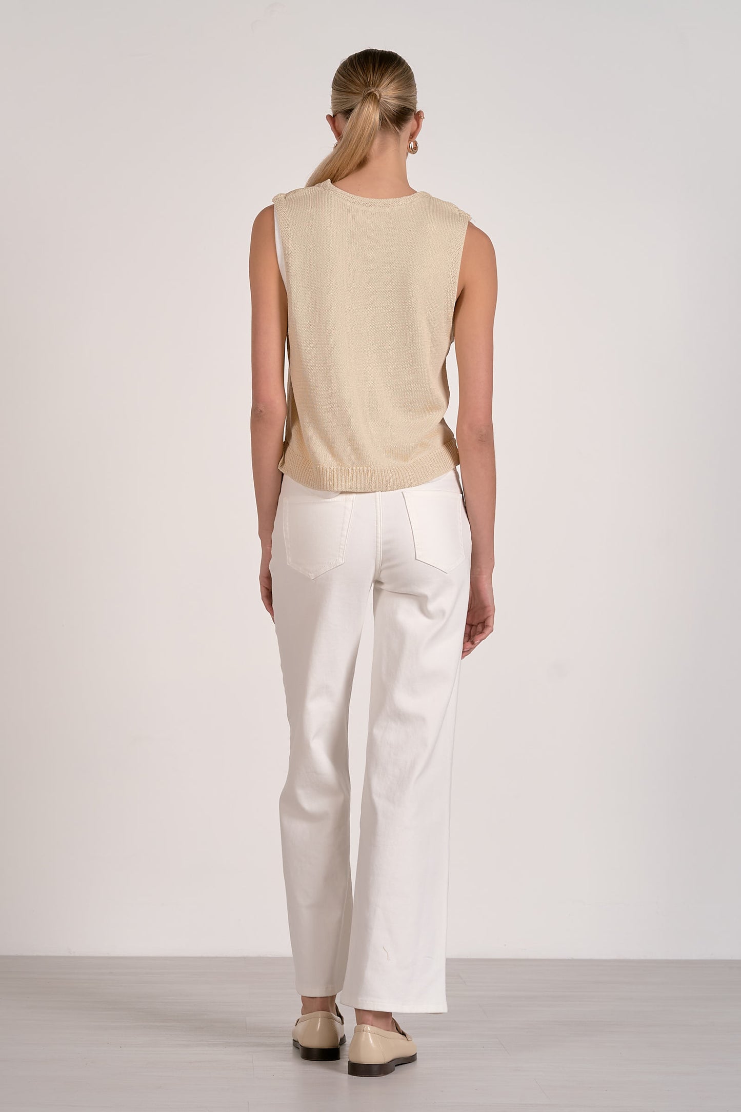 Person wearing a beige sleeveless top and white pants on a plain background