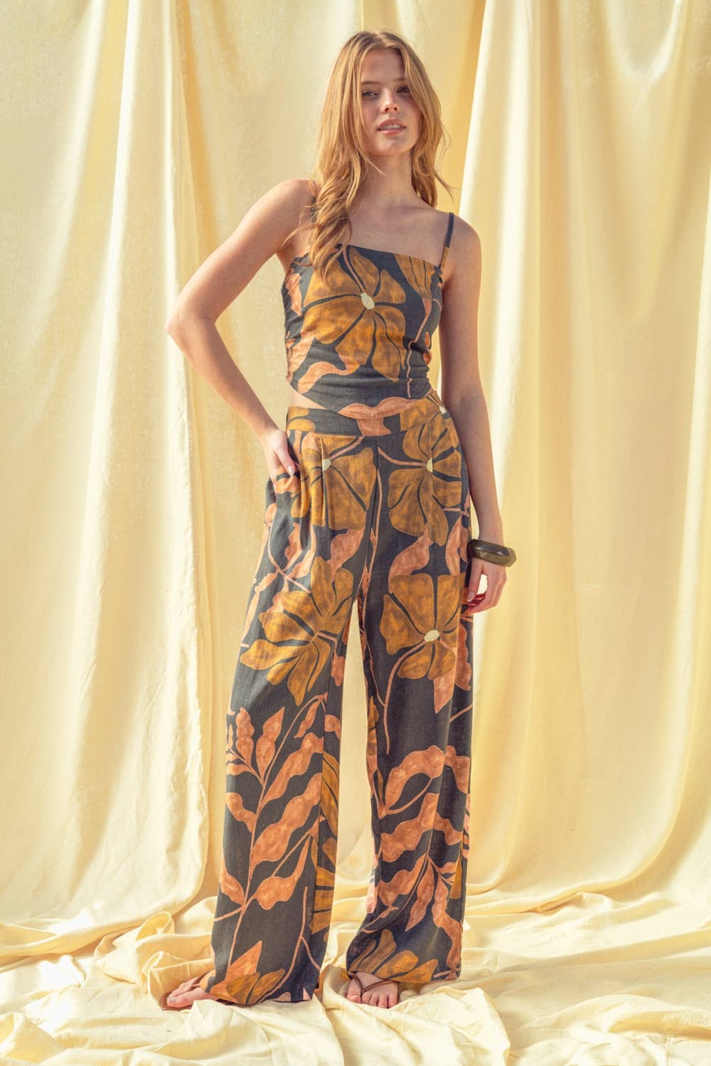 Woman wearing a floral jumpsuit standing against a beige curtain.