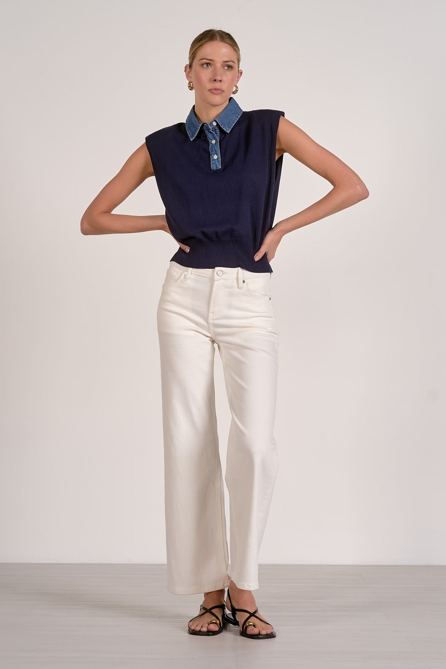 Woman wearing a navy sleeveless top with a blue collar and white pants on a plain background