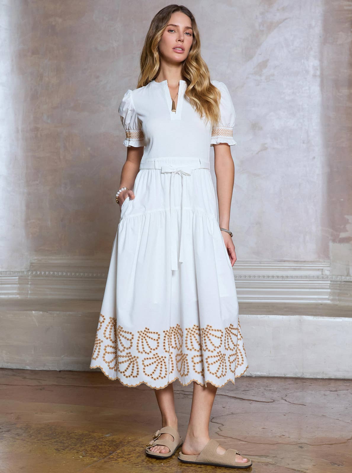 Woman wearing a white dress with lace details against a neutral wall.