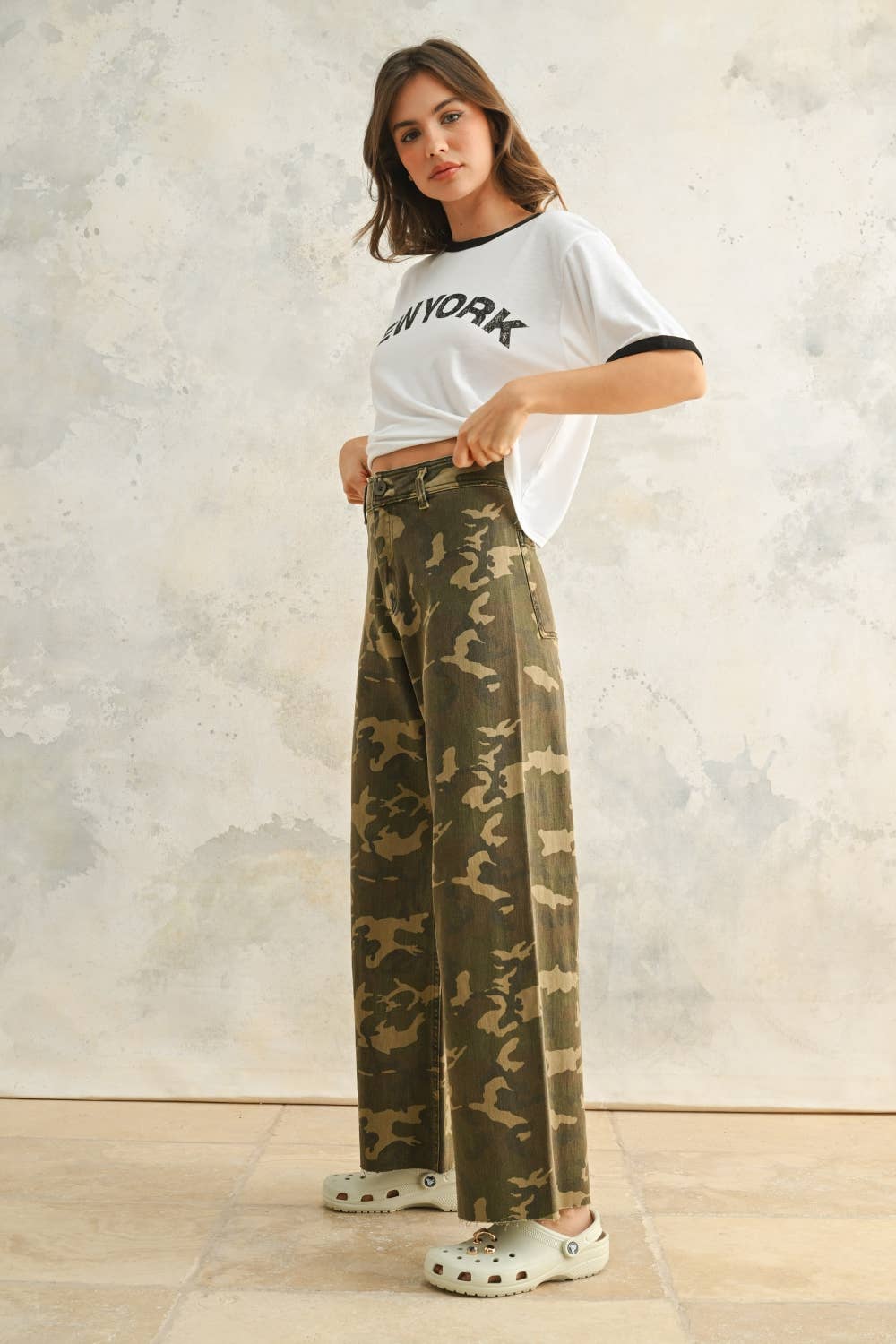 Woman wearing a white t-shirt with text and camouflage pants against a neutral background