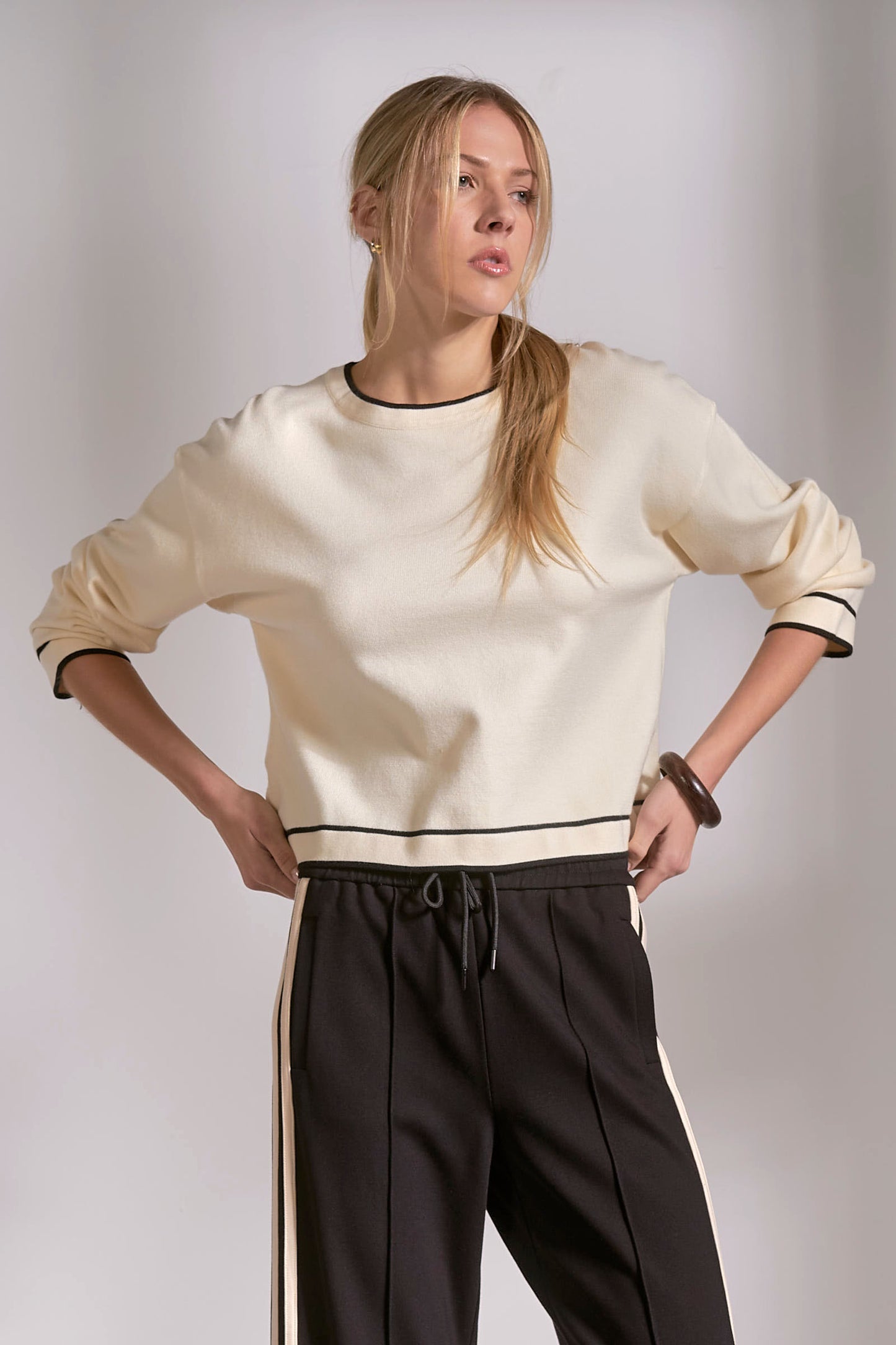 Woman wearing a cream sweater with black trim and dark pants against a plain background
