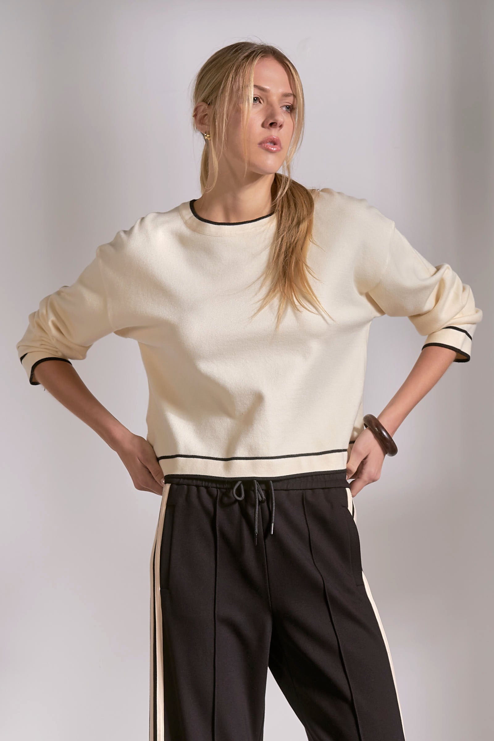 Woman wearing a cream sweater with black trim and dark pants against a plain background