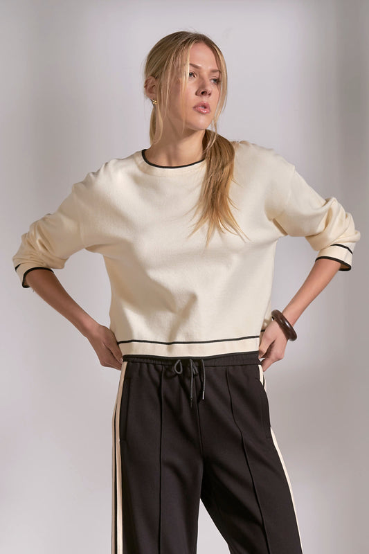 Woman wearing a cream sweater with black trim and dark pants against a plain background