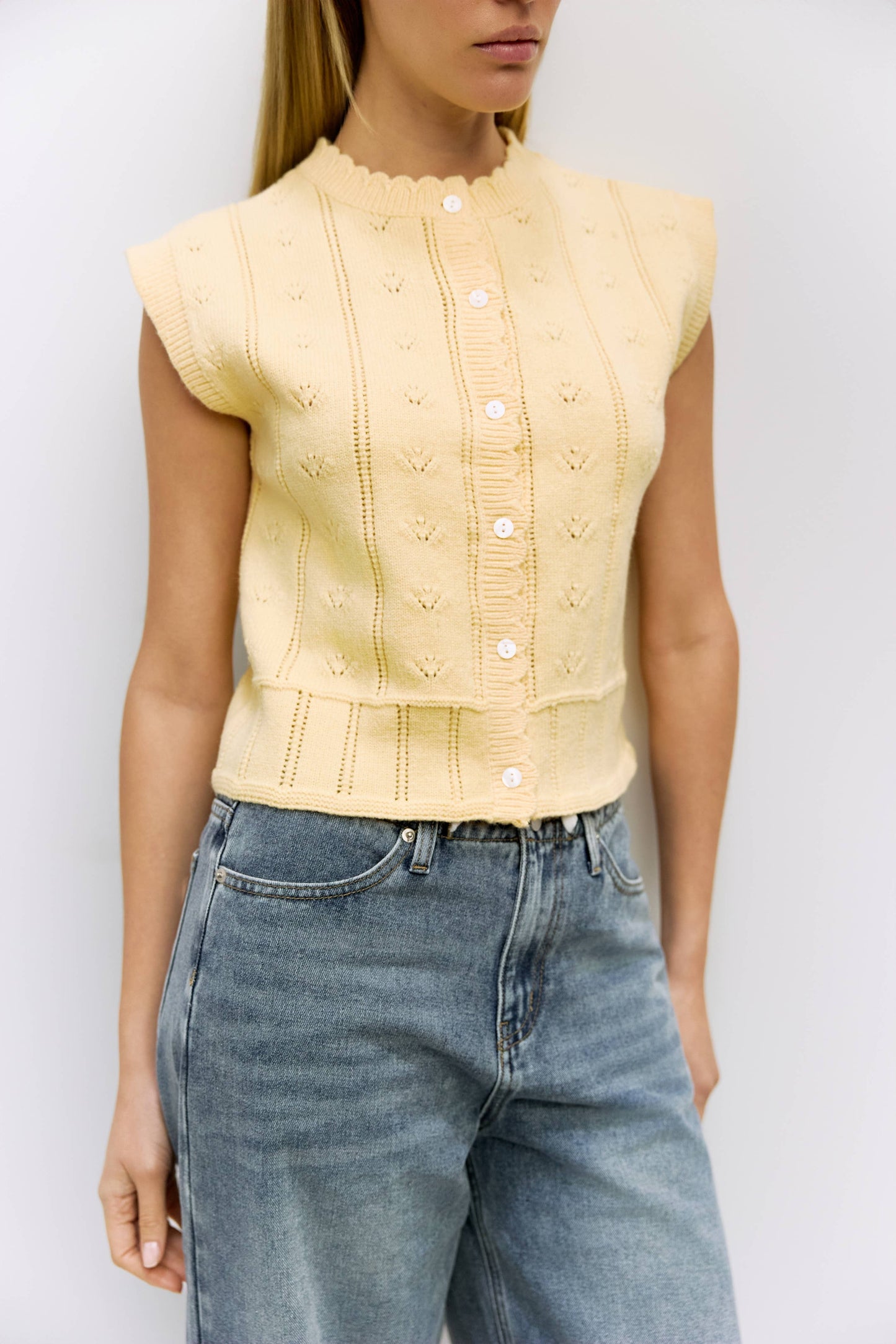 Person wearing a light yellow sleeveless top with buttons and blue jeans against a white background