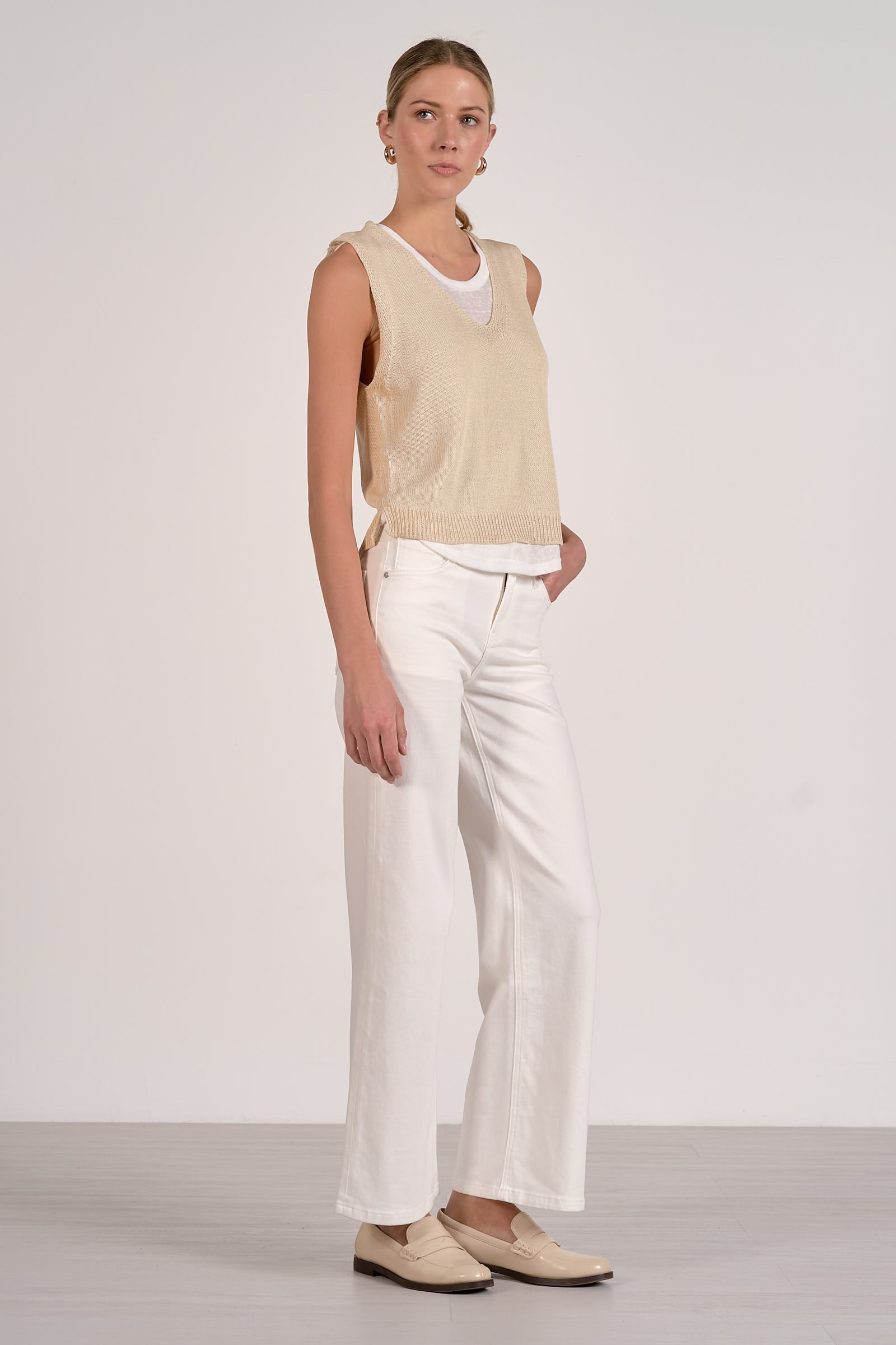 Woman wearing a beige sleeveless top and white pants against a plain background